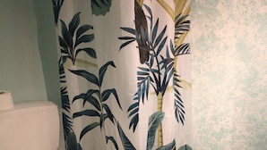A bathroom with a shower curtain featuring a tropical leaf pattern, a toilet with a green mat, and a white sink. - Alley Studio 2 by Frangki (Aegina)