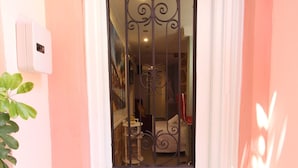 A pink exterior with a white door featuring a decorative glass panel and a gold-colored flower design below the door. - Alley Studio 2 by Frangki (Aegina)