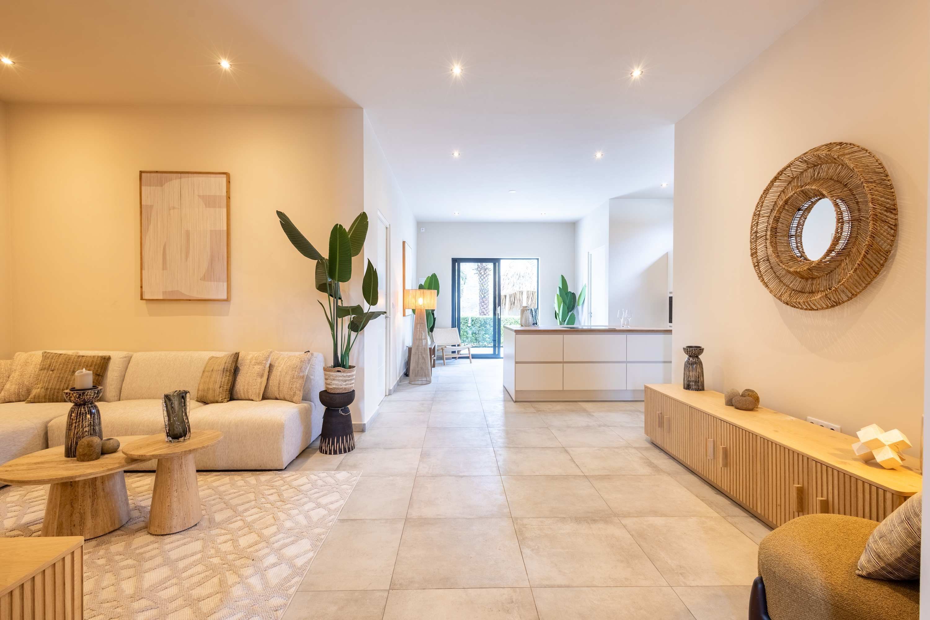 Amplia sala de estar con sofá, mesa de centro y plantas decorativas. Se ve una cocina al fondo, con encimera y armarios de madera.