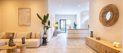 Amplia sala de estar con sofá, mesa de centro y plantas decorativas. Se ve una cocina al fondo, con encimera y armarios de madera.