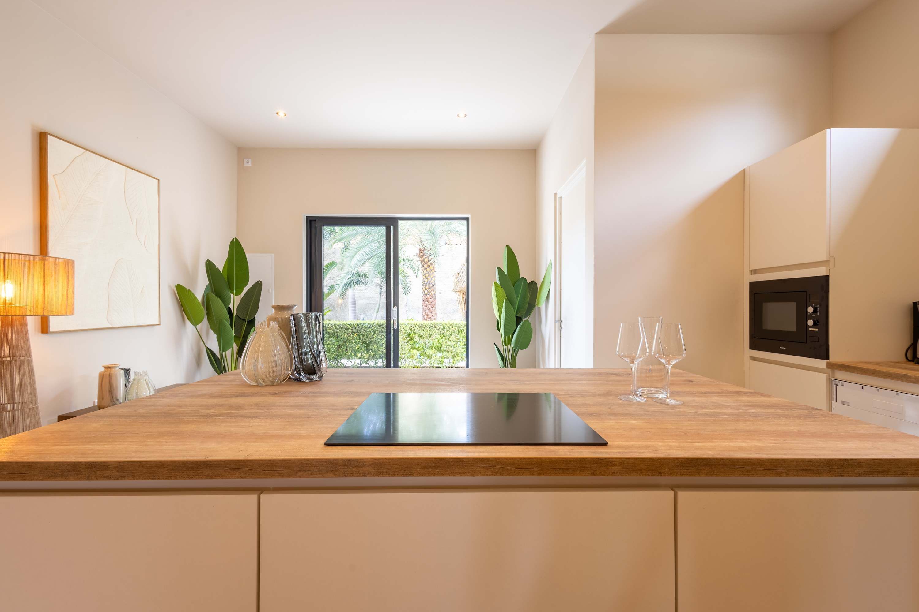 Une cuisine moderne avec un plan de travail en bois, une planche à découper noire et une vue sur les palmiers à travers la porte vitrée.