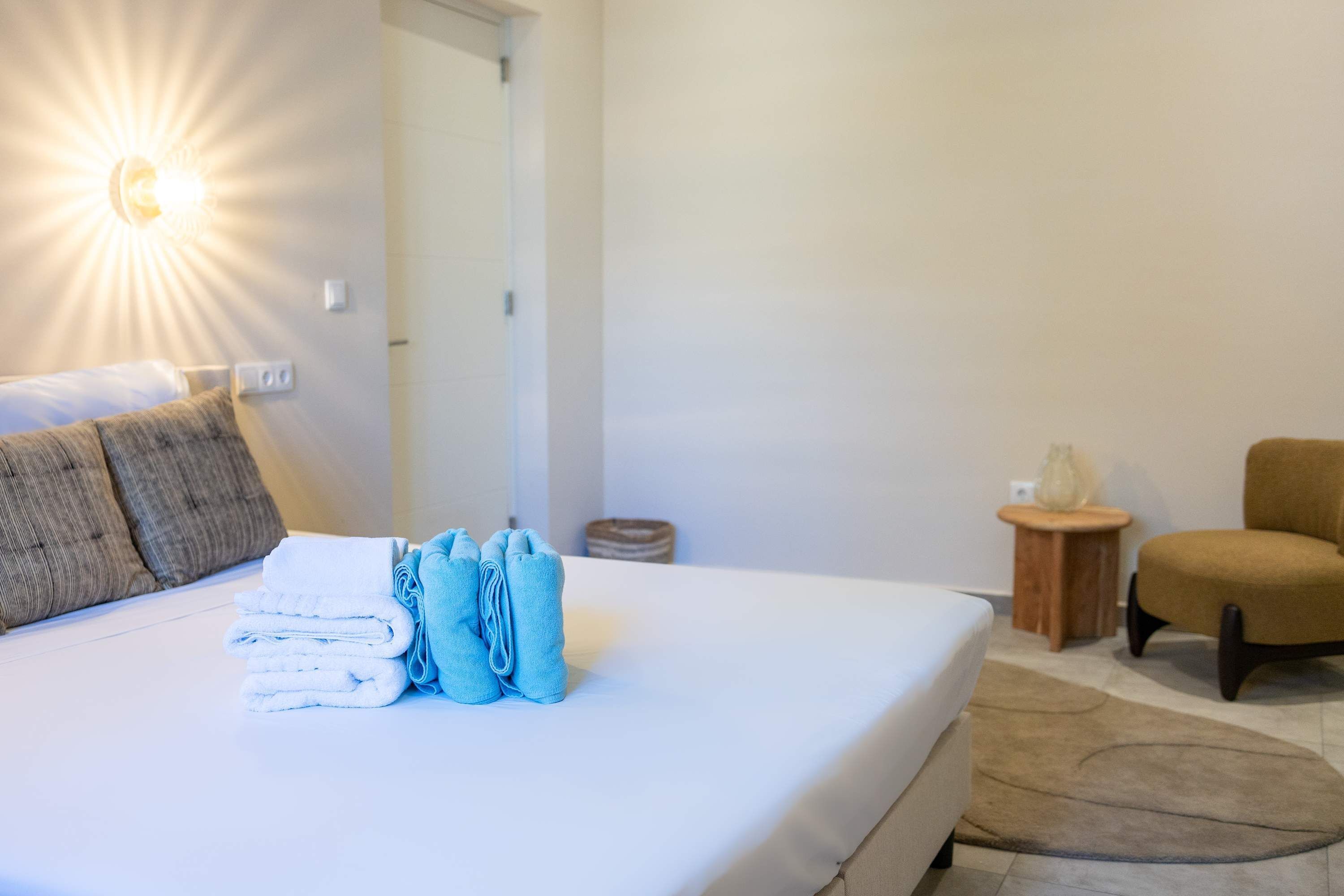 A neatly made bed with folded towels, a bedside lamp, a chair, and a small wooden side table.