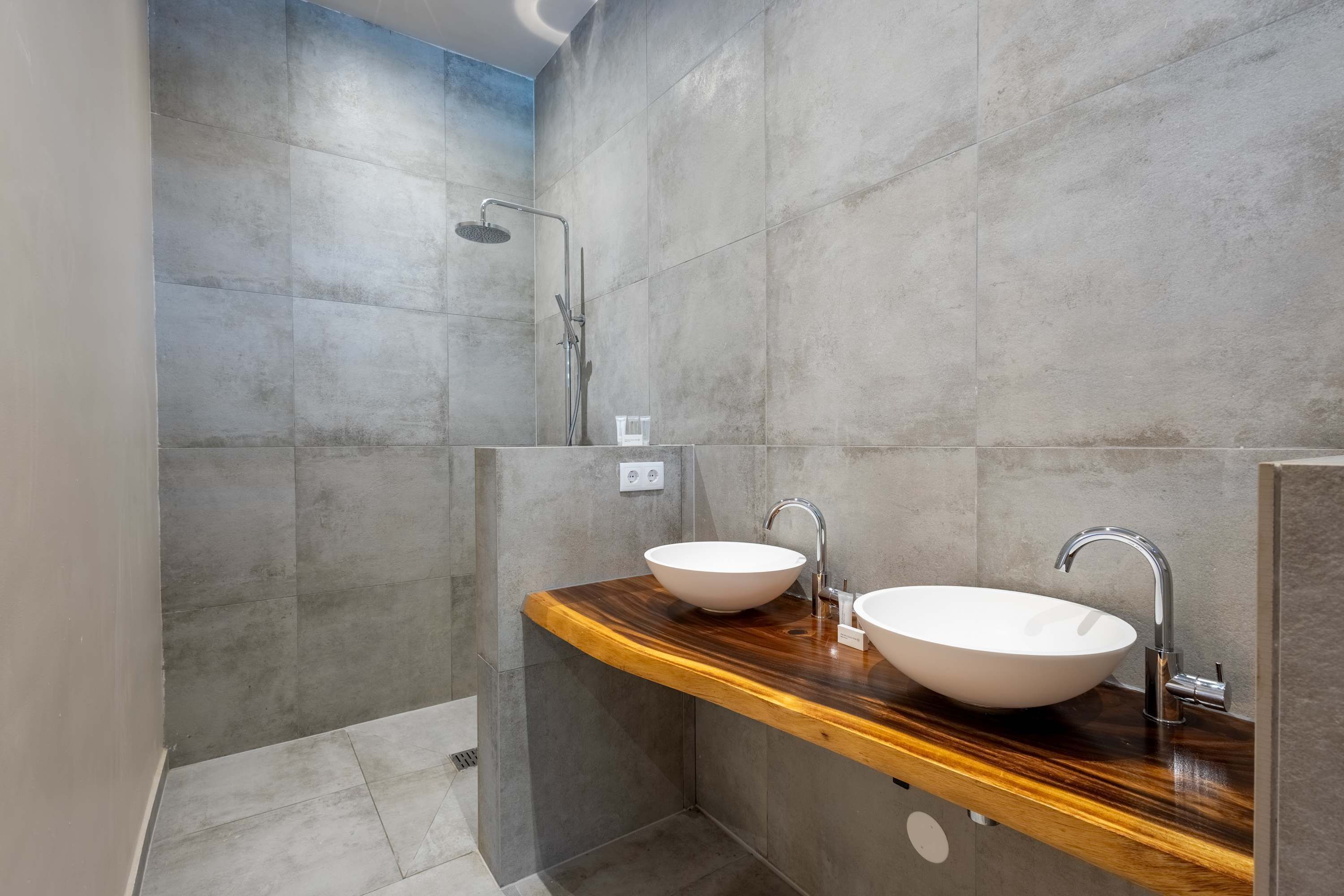 Une salle de bain moderne avec deux vasques blanches posées sur un plan de travail en bois, un espace douche et des murs recouverts de carreaux gris.