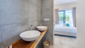 Een moderne badkamer met twee grote witte wastafels op een houten aanrecht, een spiegel en uitzicht op een slaapkamer met een bed en gordijnen.