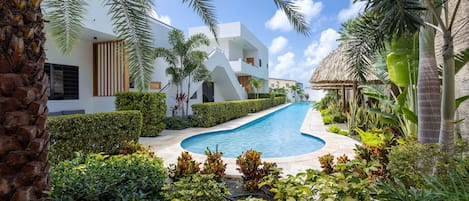 A modern resort with a swimming pool, lush greenery, and a thatched-roof structure.