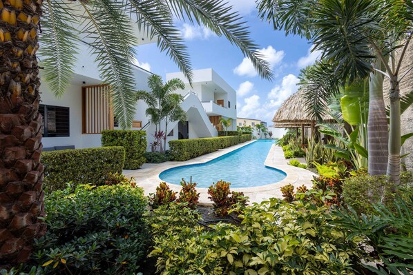 A modern resort with a swimming pool, lush greenery, and a thatched-roof structure.