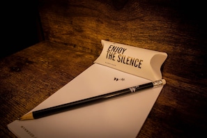 Une table en bois avec un carnet blanc et un crayon. Sur le carnet, on peut lire les mots « PROFITEZ DU SILENCE » et « BOULES QUIES ».