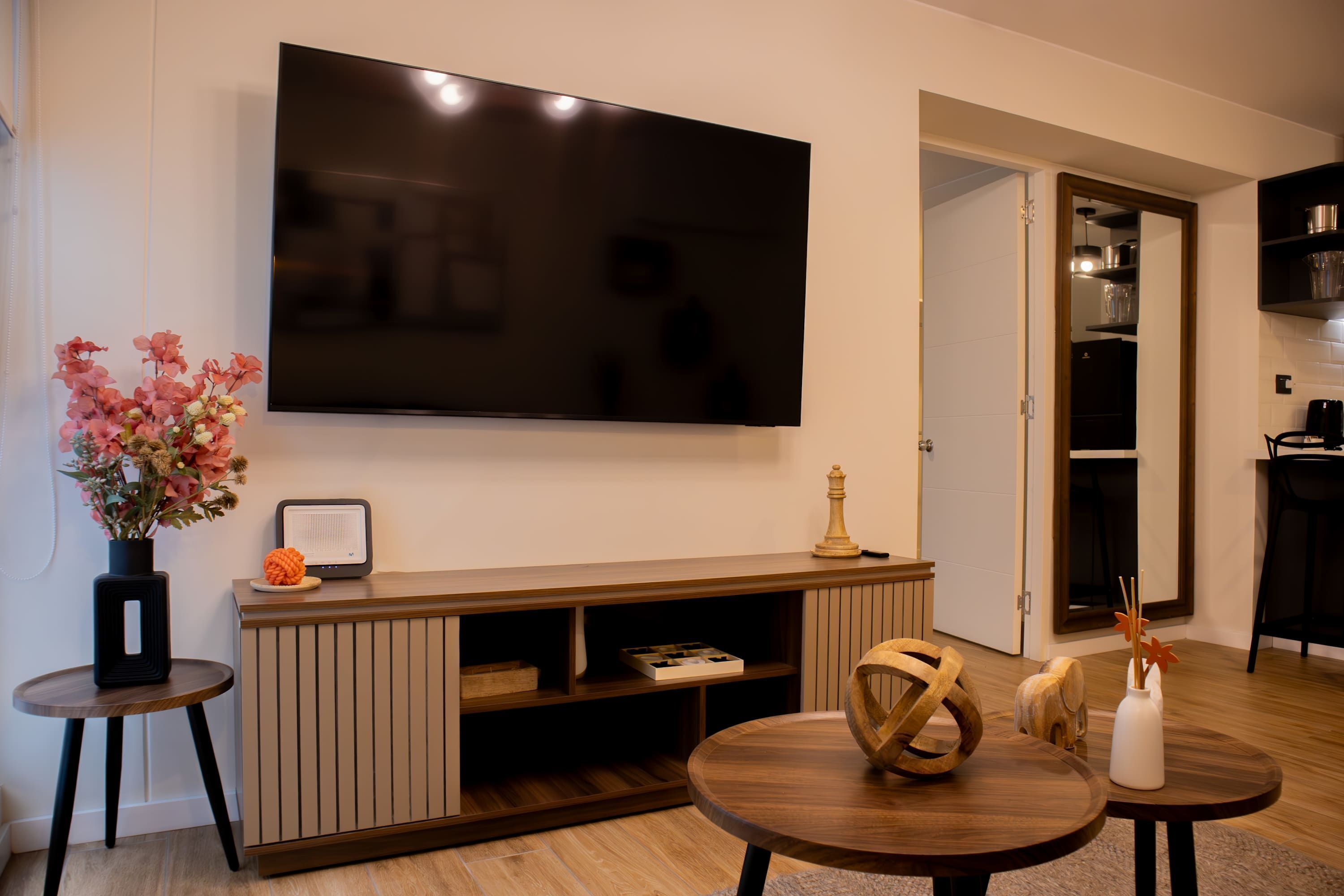 A modern living room with a flat-screen TV mounted on the wall, a wooden entertainment unit, two round wooden tables, and a small side table with a vase of flowers.