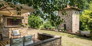 Un edificio de piedra con una pequeña torre, rodeado de vegetación y un patio con mobiliario exterior.