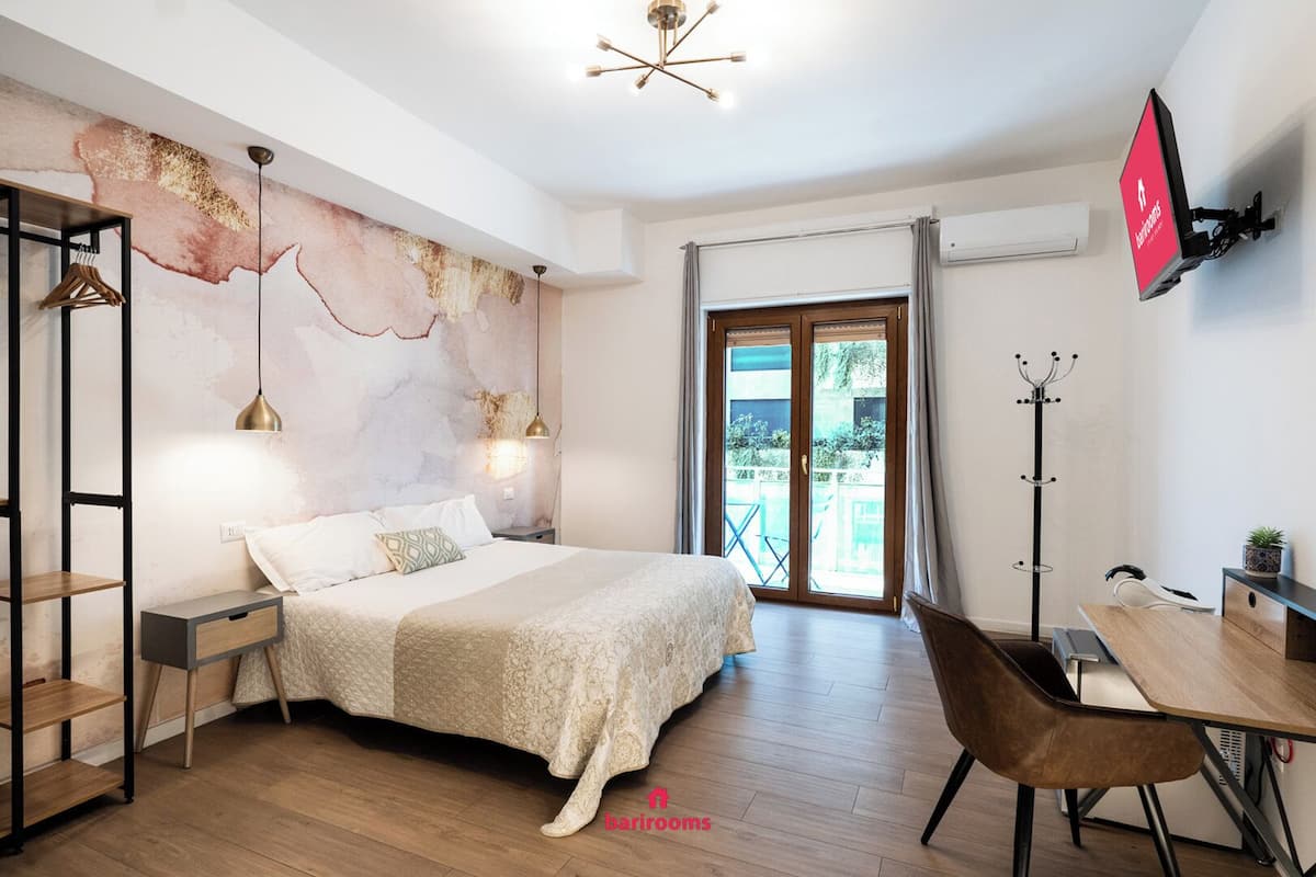 A modern bedroom with a bed, bedside tables, a desk, and a chair. There is a large window with curtains and a view of greenery outside.