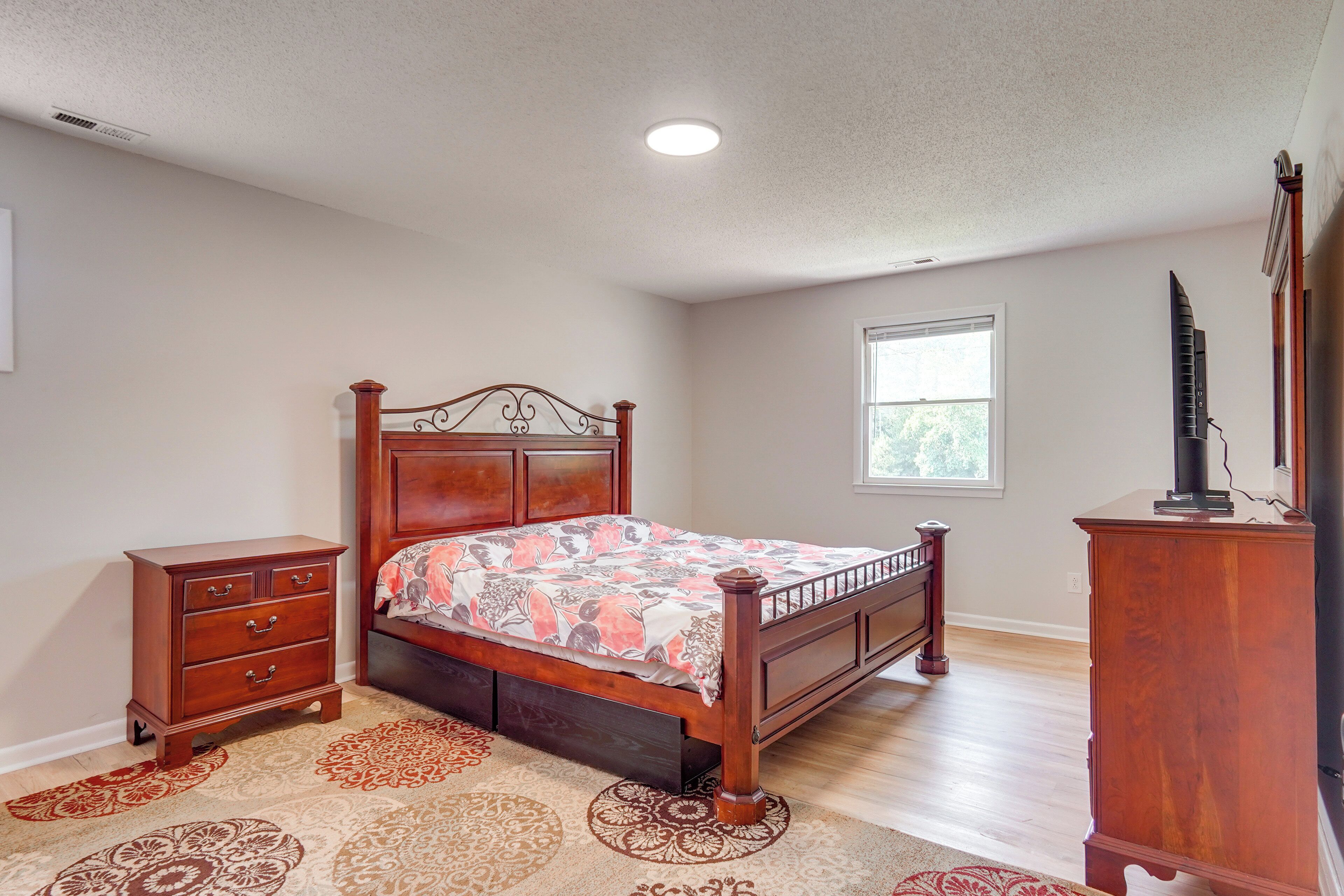 Une chambre à coucher avec un lit en bois, une table de chevet, une commode et une fenêtre laissant entrer la lumière naturelle.