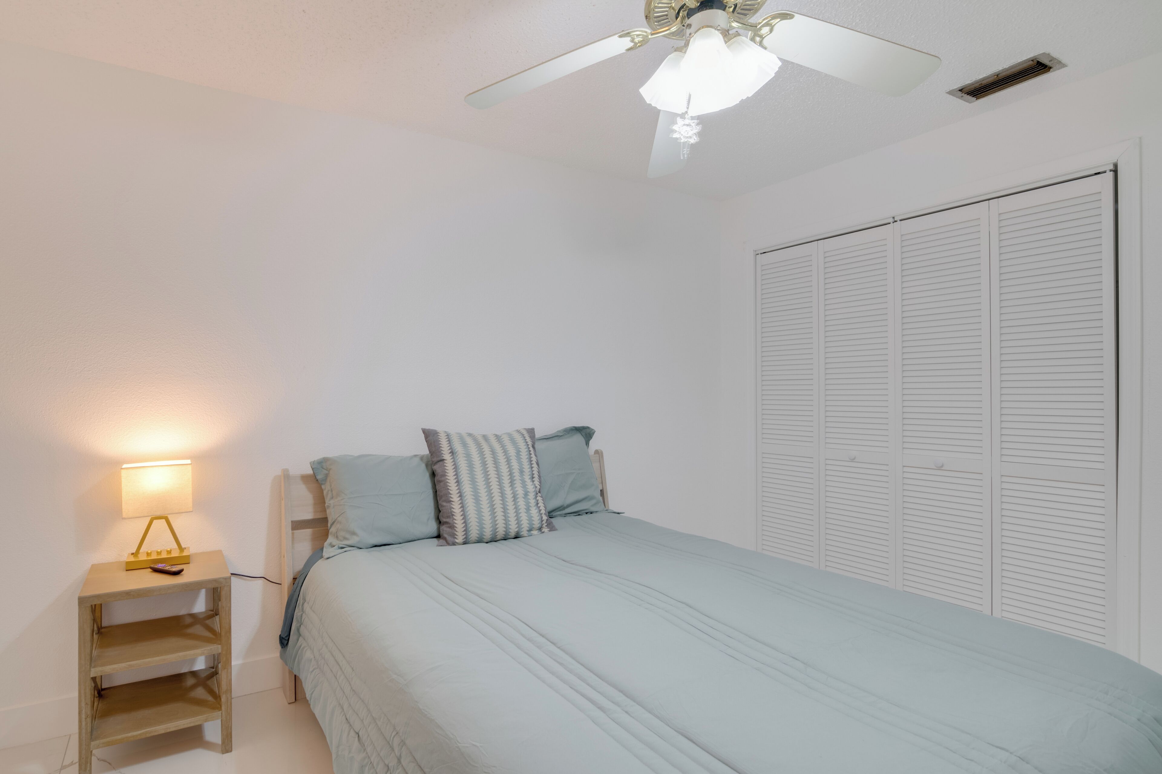 A bedroom with a bed, a nightstand with a lamp, a ceiling fan, and a wardrobe.