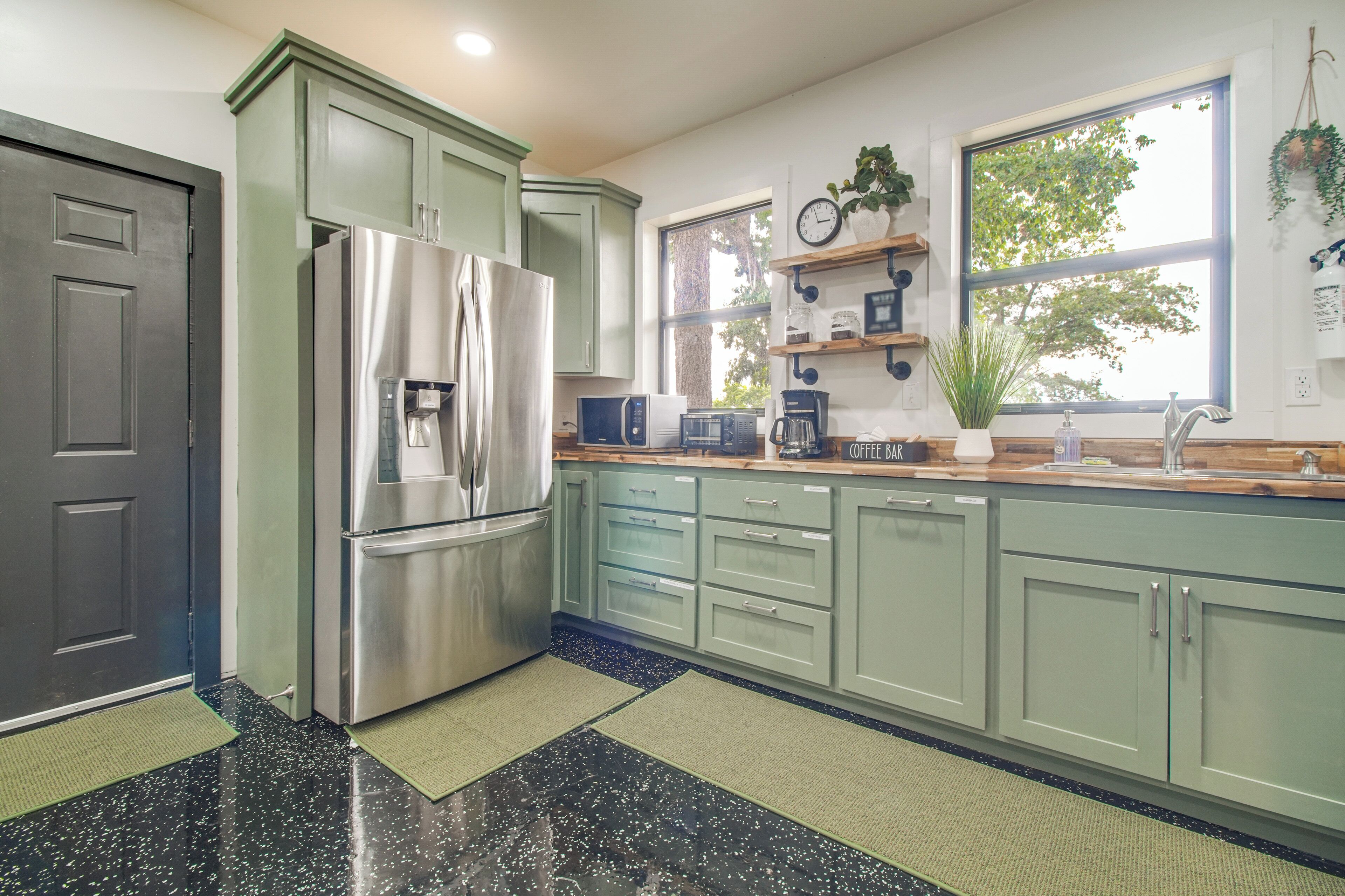 A kitchen with green cabinets, a stainless steel refrigerator, a coffee maker, and a window with a view of trees.