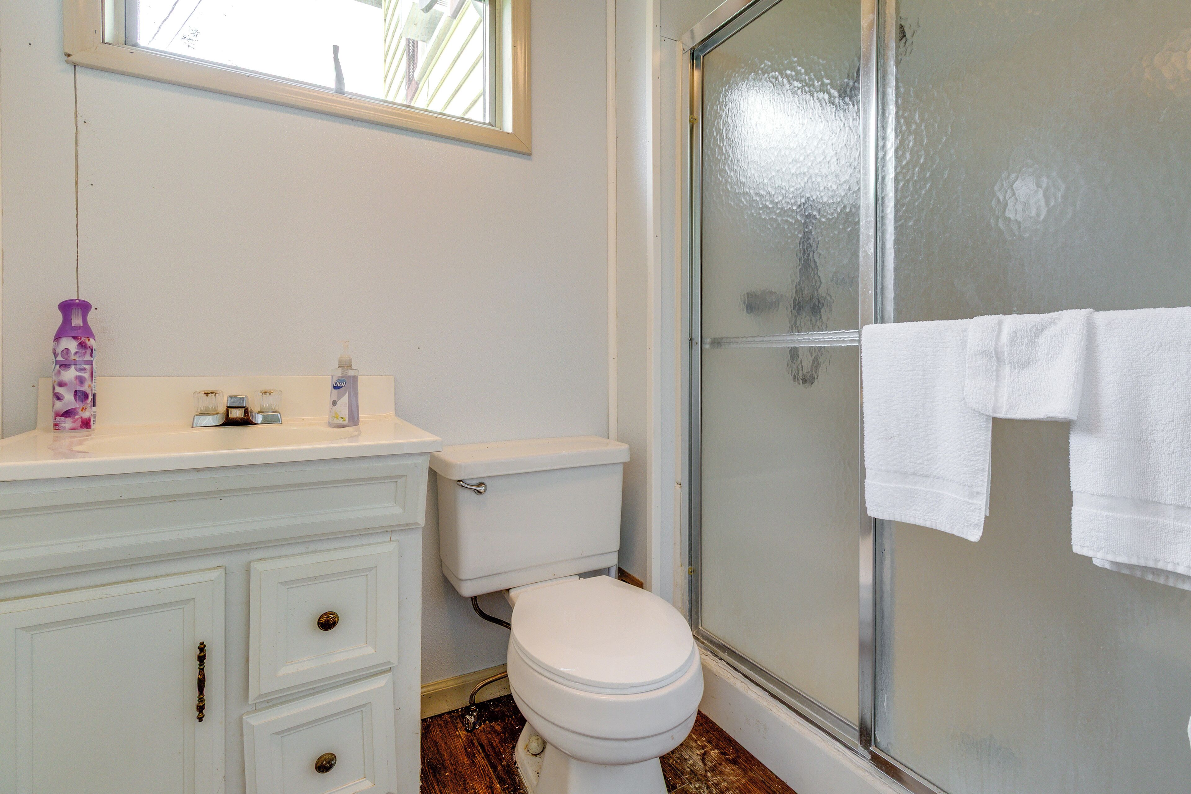 A bathroom with a white toilet, a vanity with drawers, a window, and a glass shower enclosure.