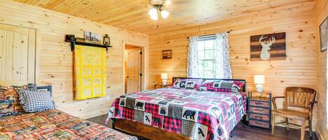 A wooden cabin bedroom with a large bed, a smaller bed, a chair, a desk, and a ceiling fan.