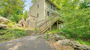 A two-story house with a wooden staircase leading to the entrance, surrounded by lush greenery and a stone wall. - Walk to Putnam Lake: Updated Home w/ Deck (Patterson)
