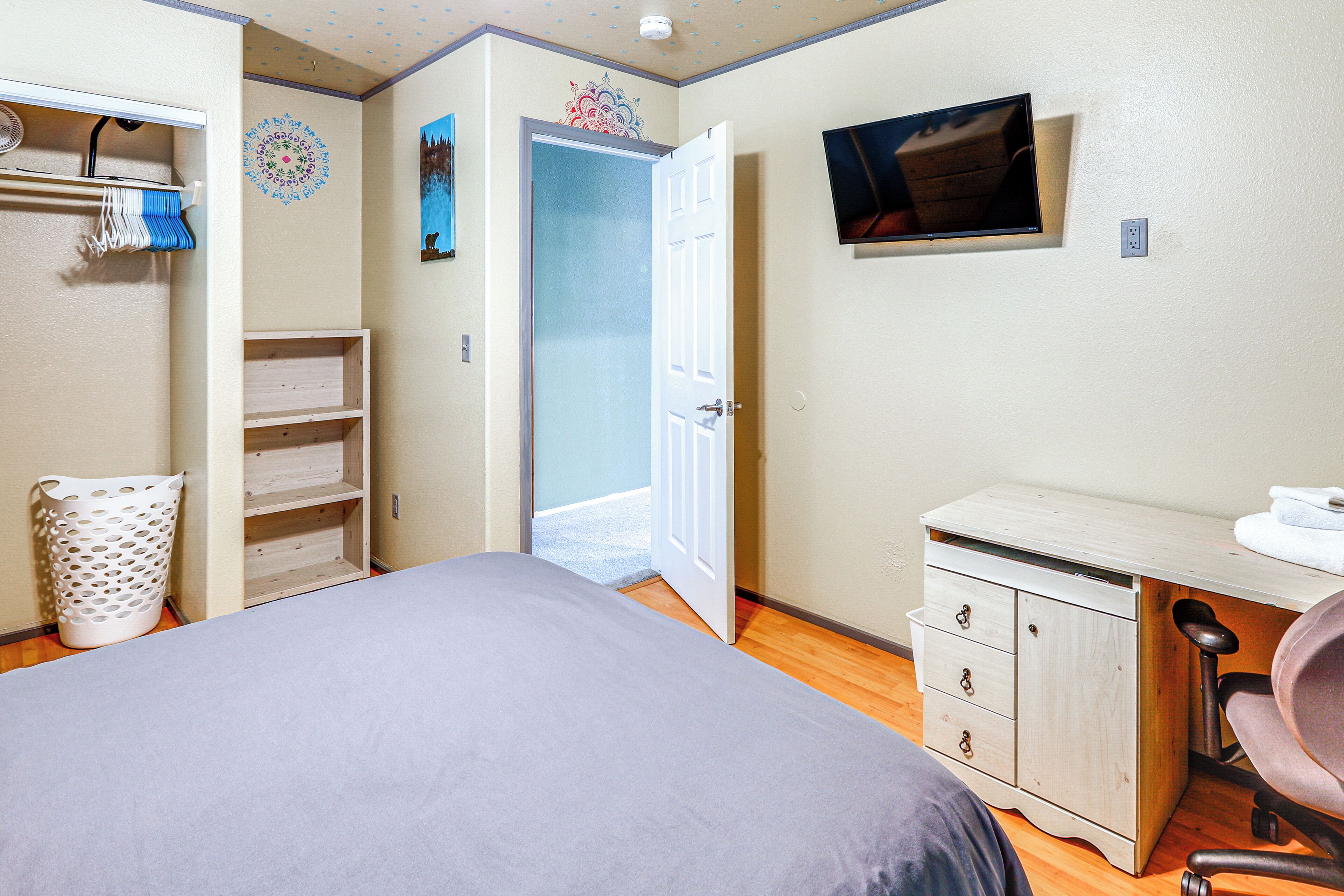 A bedroom with a bed, a desk, a chair, a TV, and a door leading to another room.