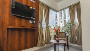 Una habitación con televisión montada en una pared de madera, una mesita con un florero, una silla y una ventana con cortinas.