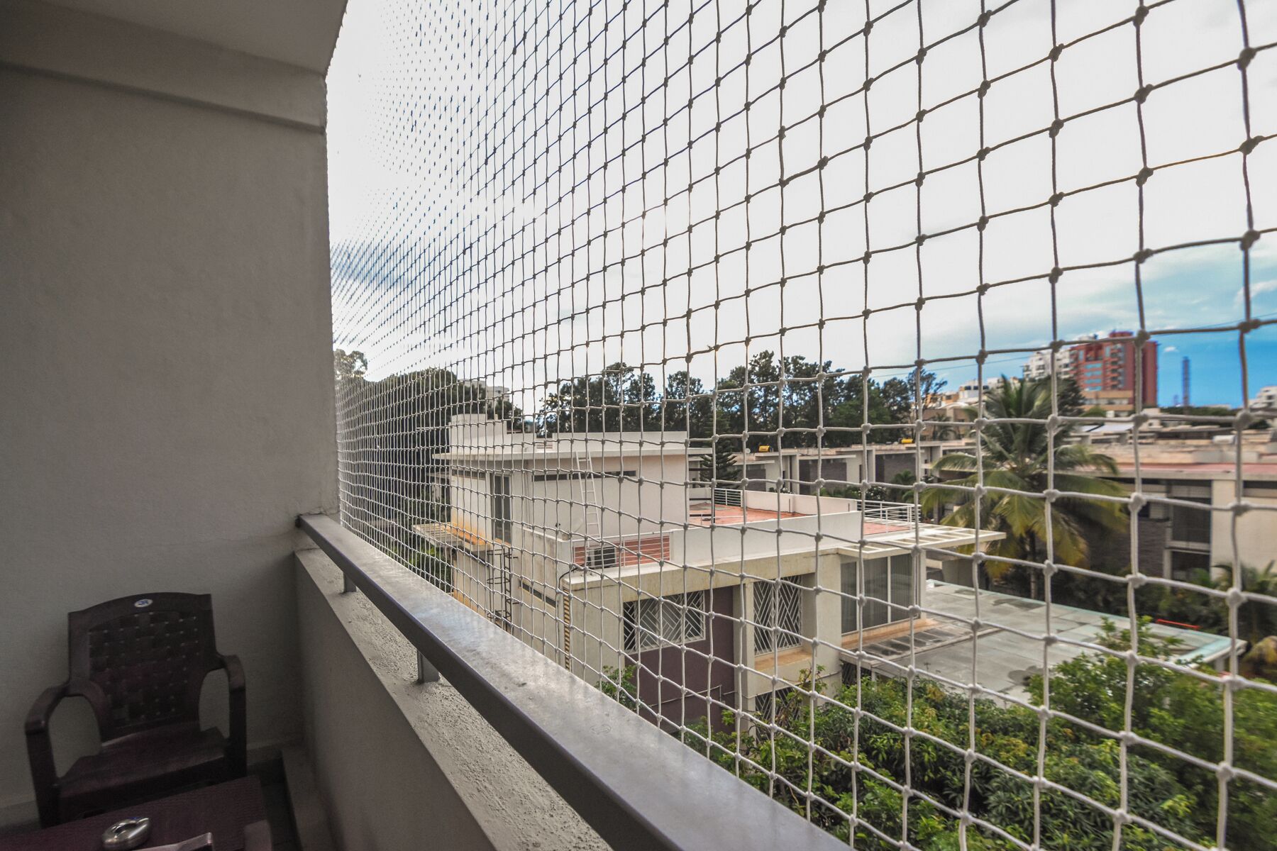 A balcony with a metal safety screen, a chair, and a view of buildings and greenery.