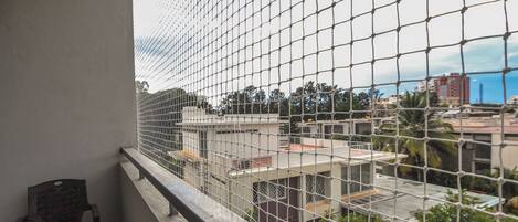 A balcony with a metal safety screen, a chair, and a view of buildings and greenery.