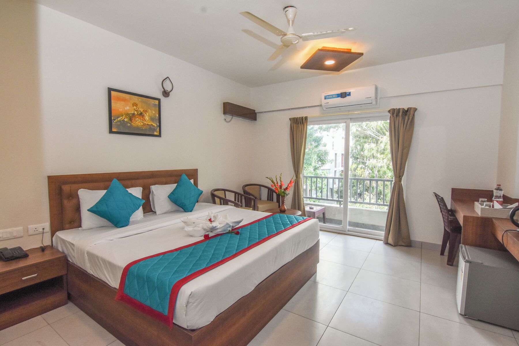 A hotel room with a bed, a desk, a chair, a ceiling fan, an air conditioning unit, a window with curtains, and a painting on the wall.