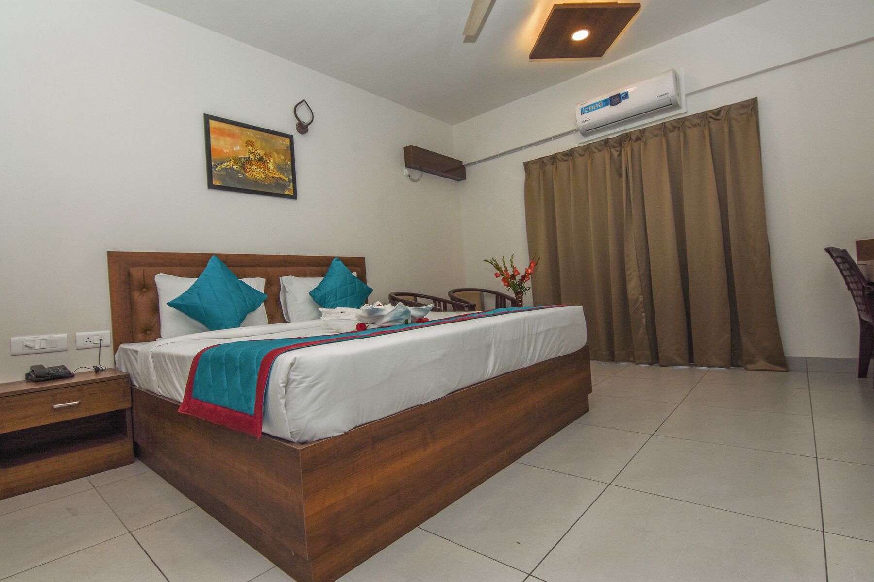 Ein Hotelzimmer mit einem hölzernen Bett, weißer Bettwäsche, türkisfarbenen Kissen, einem Gemälde an der Wand, einer Klimaanlage und einem Fenster mit Vorhängen.