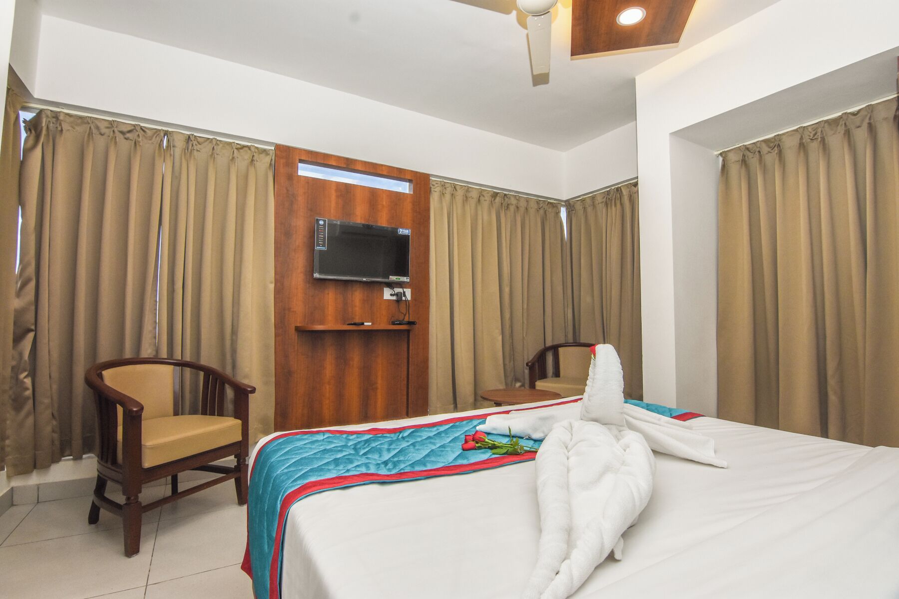A hotel room with a bed, two chairs, a TV mounted on the wall, and curtains.