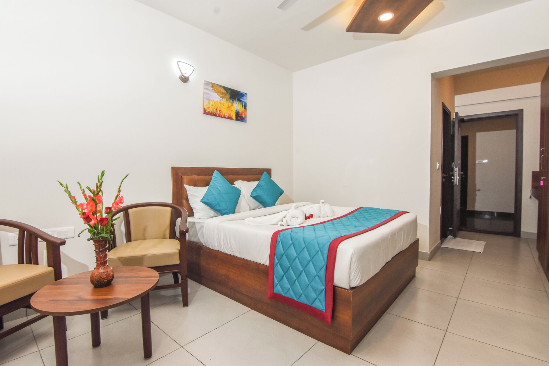 A hotel room with a bed, a chair, a small table, and a vase of flowers.