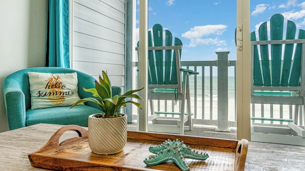 Un canapé de couleur turquoise agrémenté d’un coussin orné de l’inscription « bonjour, été », une plante en pot, un plateau en bois avec une étoile de mer, et une vue sur une plage avec des chaises Adirondack vertes, visible à travers une porte vitrée.