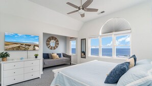 Une chambre à coucher comprenant un lit, une commode, un canapé, un ventilateur de plafond et une grande fenêtre donnant sur l’océan.