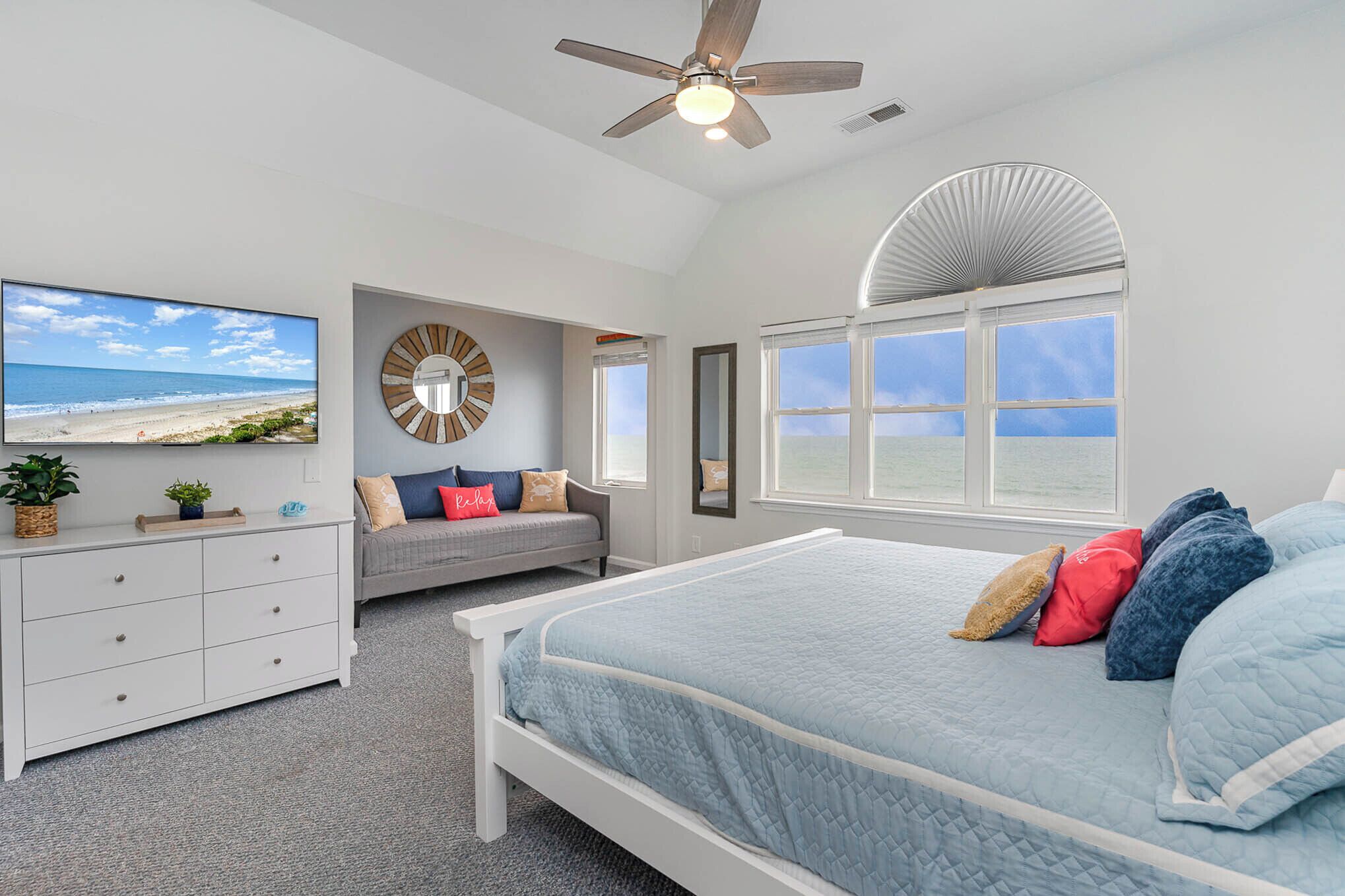 A bedroom with a bed, a TV, a ceiling fan, a window, and a view of the beach.