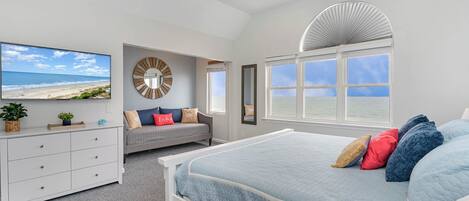 A bedroom with a bed, a TV, a ceiling fan, a window, and a view of the beach.