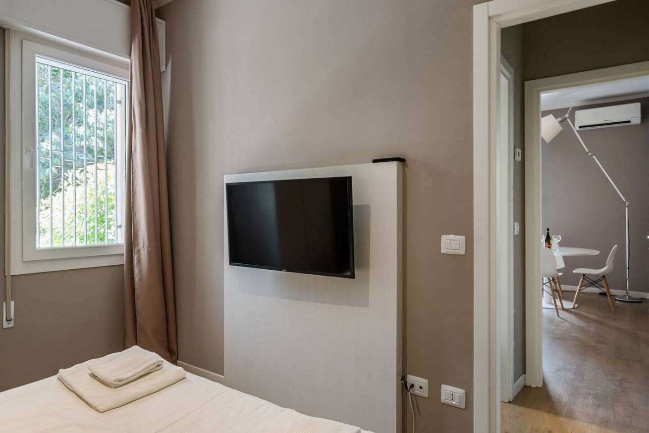 Ein Hotelzimmer mit Bett, einem an der Wand montierten Fernseher und einem Fenster mit Vorhängen.