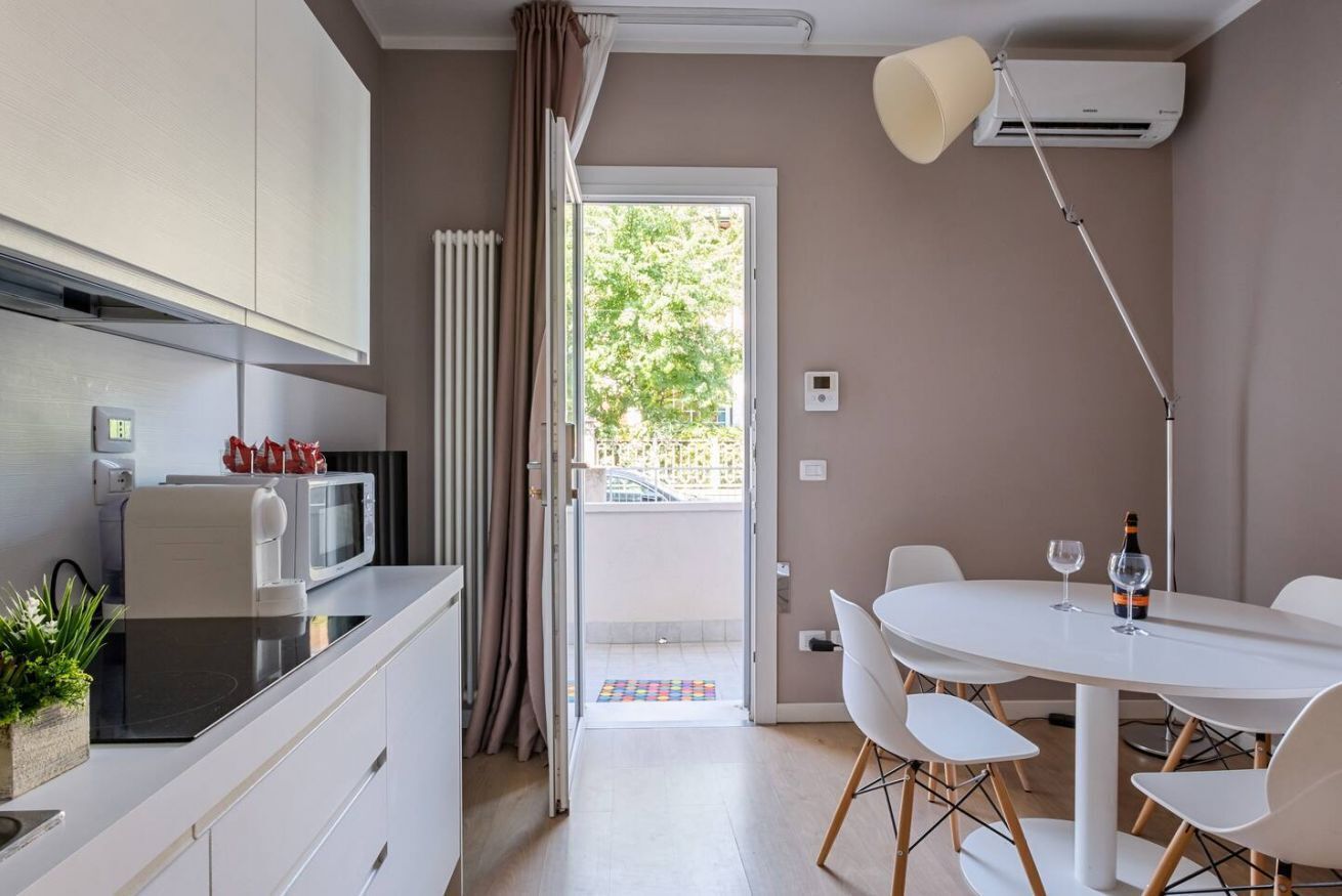 A modern kitchen with a microwave, toaster, and a plant on the counter. There is a dining area with a white table and chairs, and a ceiling lamp.