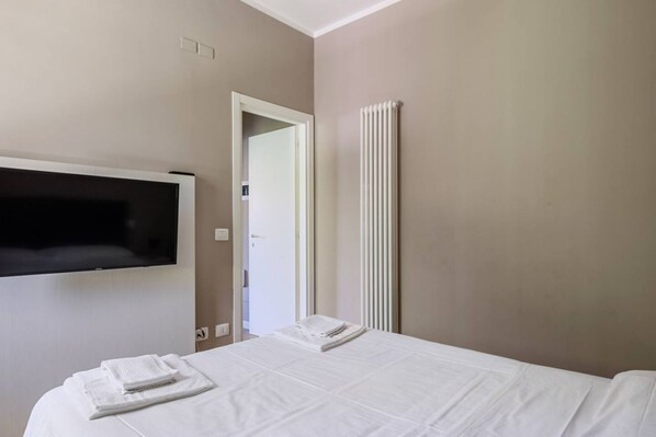 Habitación de hotel con cama, televisor montado en la pared, radiador y puerta que da a otra habitación.