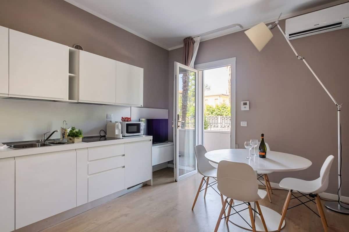 Eine moderne Küche mit weißen Schränken, einer Mikrowelle und einem Spülbecken. Es gibt einen Essbereich mit einem weißen Tisch und Stühlen, sowie einen Balkon mit Blick.
