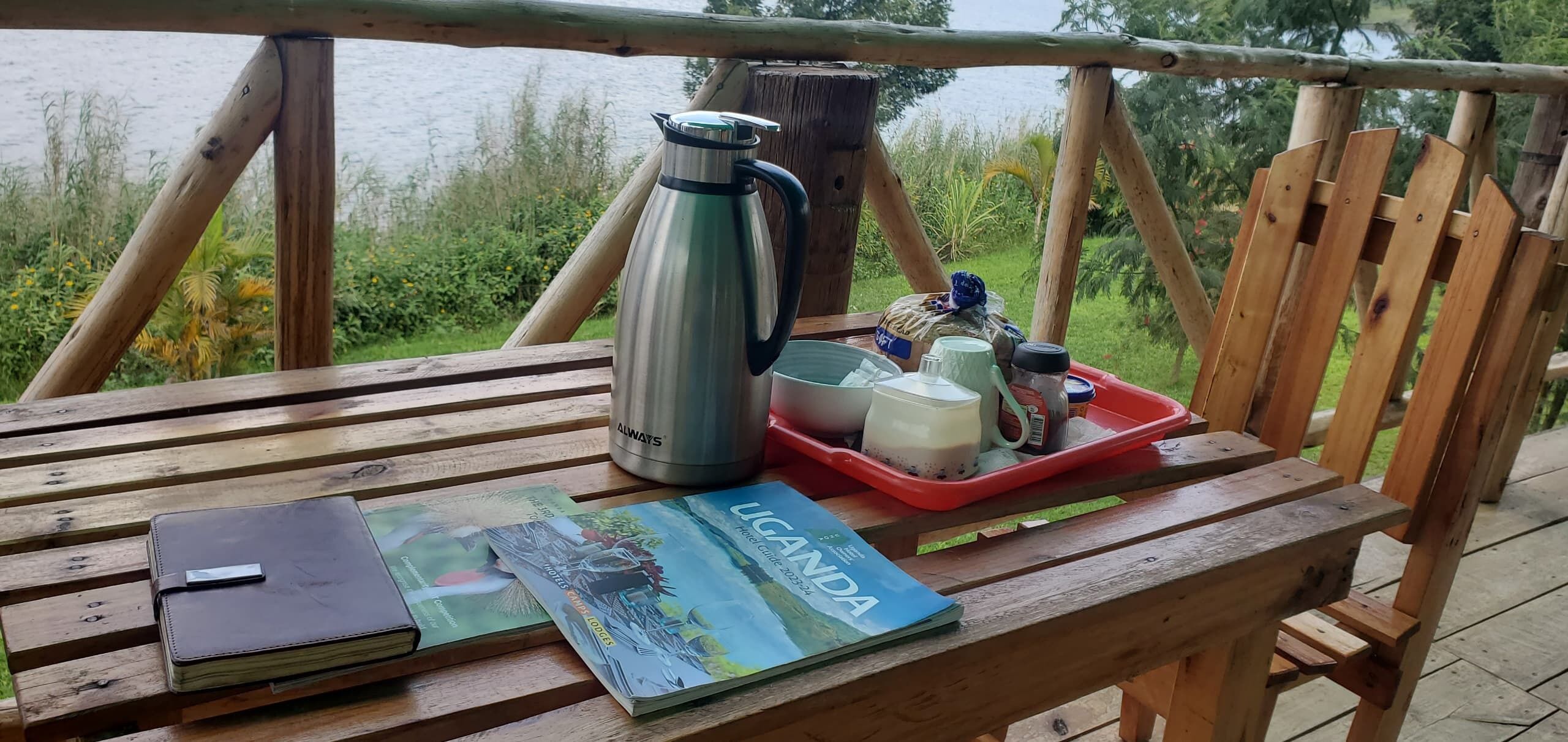 A wooden table with a travel magazine, a notebook, a stainless steel thermos, a red tray with a mug and condiments, and a wooden chair.