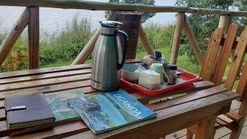 A wooden table with a travel magazine, a notebook, a stainless steel thermos, a red tray with a mug and condiments, and a wooden chair.