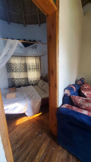 Quarto com cama sob mosquiteiro, piso de madeira e sofá com almofadas estampadas.