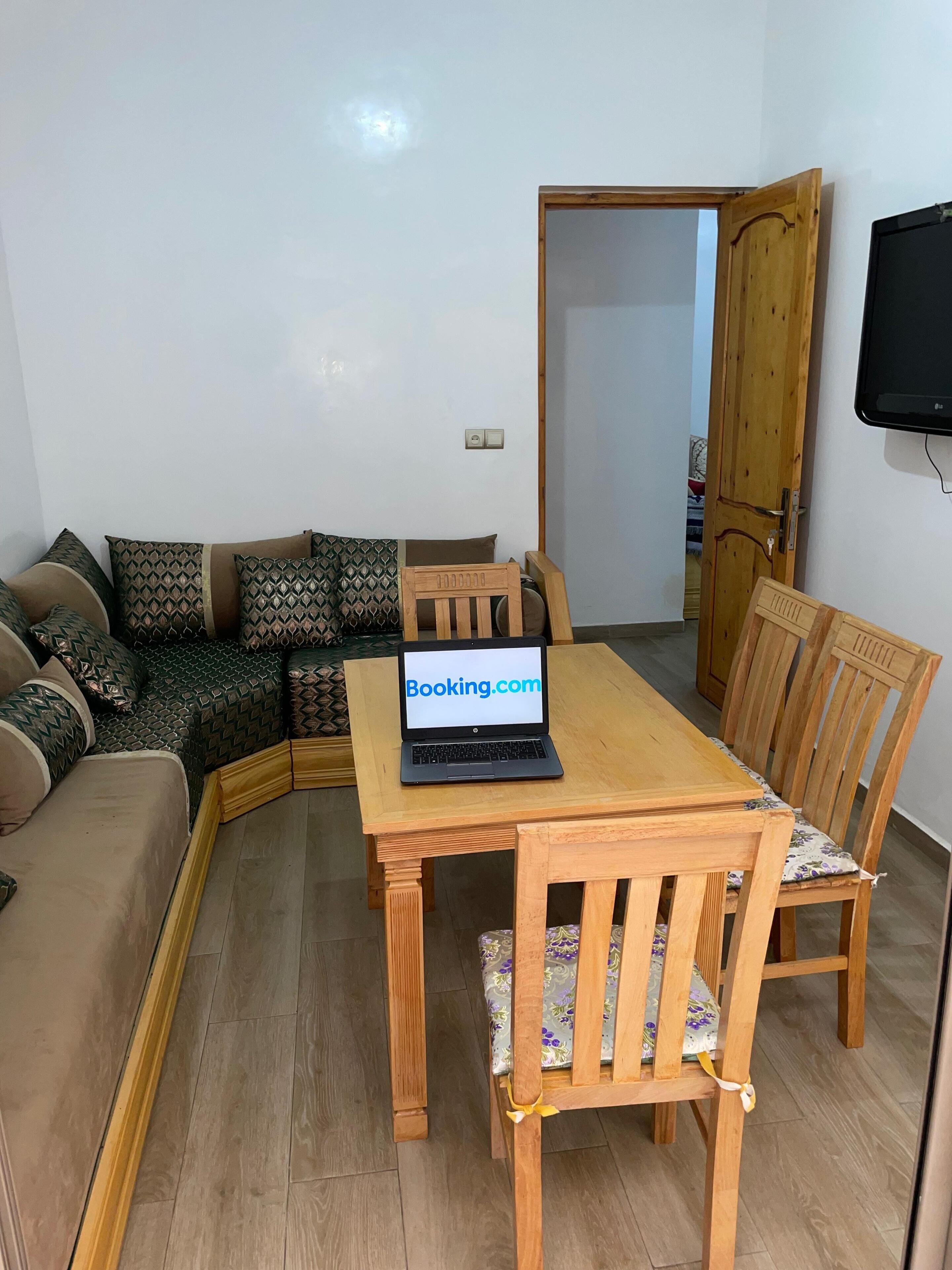 Un salon avec une table à manger en bois, un canapé agrémenté de coussins à motifs et un ordinateur portable affichant le site « Booking.com ».