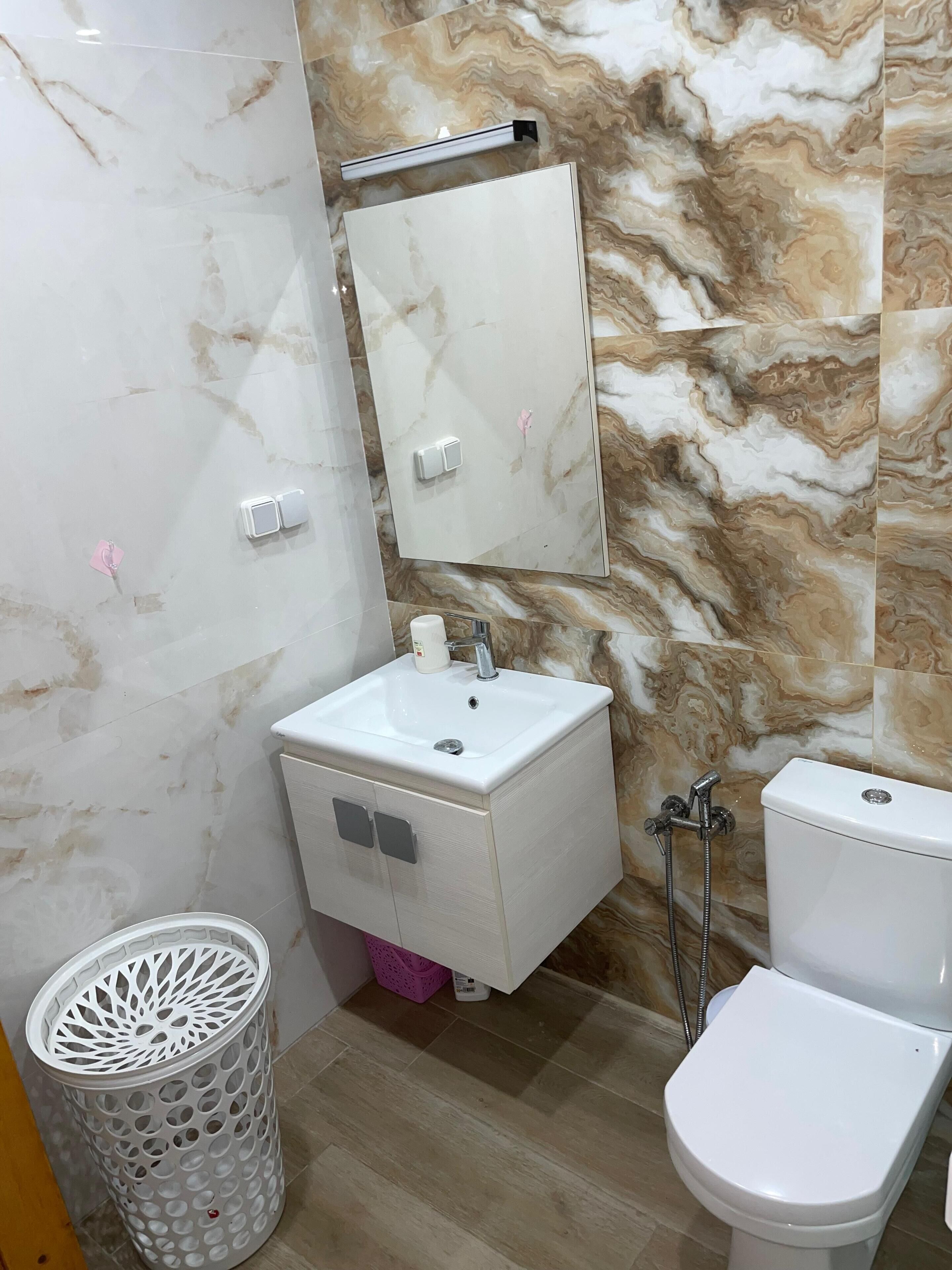 Une salle de bain avec un pare-baignoire en marbre, un lavabo sur colonne, des toilettes et une poubelle.
