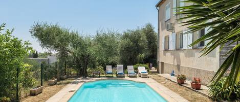 Ein rechteckiger Pool mit einer gefliesten Kante, umgeben von einer Betonplattform. Drei Liegestühle mit blauen Kissen reihen sich entlang des Poolrandes. Das Pool-Areal grenzt an ein Gebäude mit beige-farbener Fassade und weißen Fensterläden.