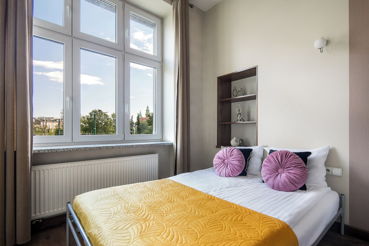 Un dormitorio con cama, ventana con vista a edificios, radiador y estante con adornos.