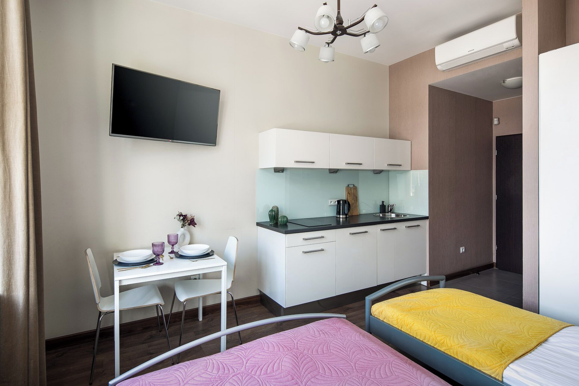 A compact hotel room with a kitchenette, a bed with a yellow mattress, a desk with a chair, and a TV mounted on the wall.