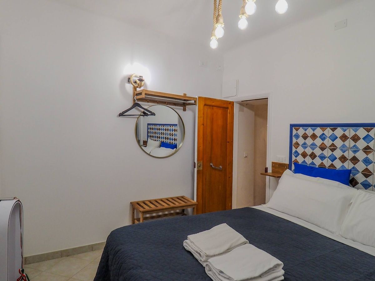 Un dormitorio con cama, mesita de noche, lámpara colgante, espejo y puerta.