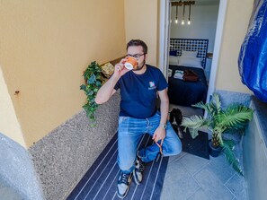 Un homme est assis sur une marche à l’extérieur d’un bâtiment et tient une tasse à la main. Un chien se trouve à proximité, et la zone est agrémentée de plantes et d’une bâche bleue.