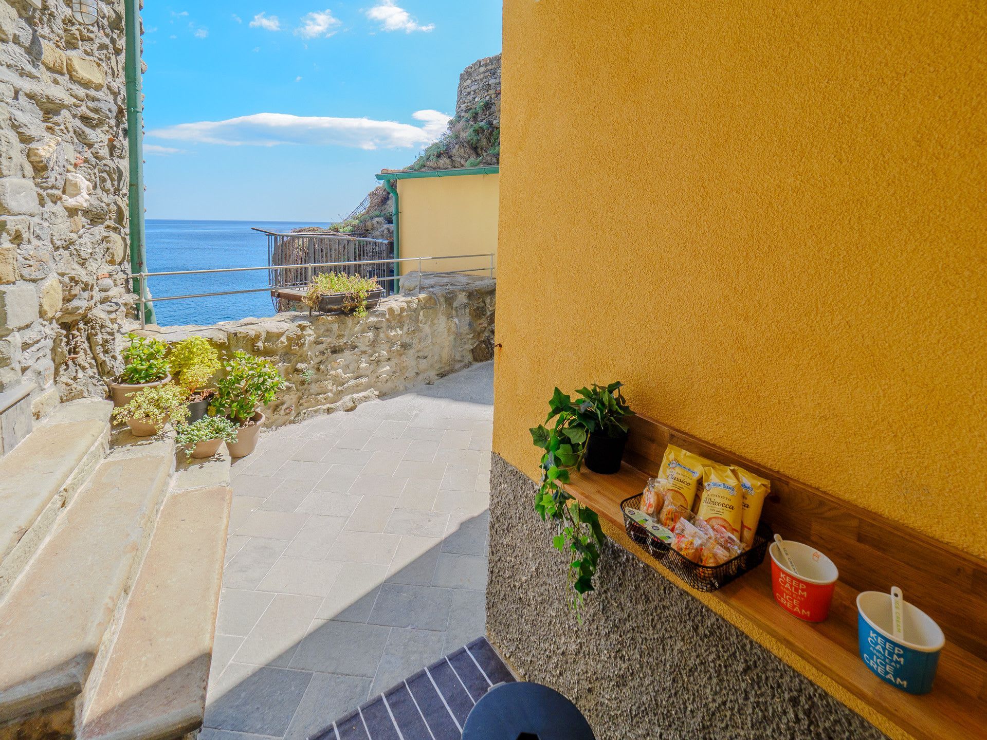 A stone staircase leading to a balcony overlooking the sea, with potted plants and a shelf with snacks and a red mug.