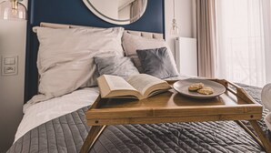 A neatly made bed with a quilted blanket, a bedside table with a tray holding an open book and cookies, and a round mirror on the wall. - Rosa - Obud si w Sercu pi Knej Bydgoszczy (Bydgoszcz)