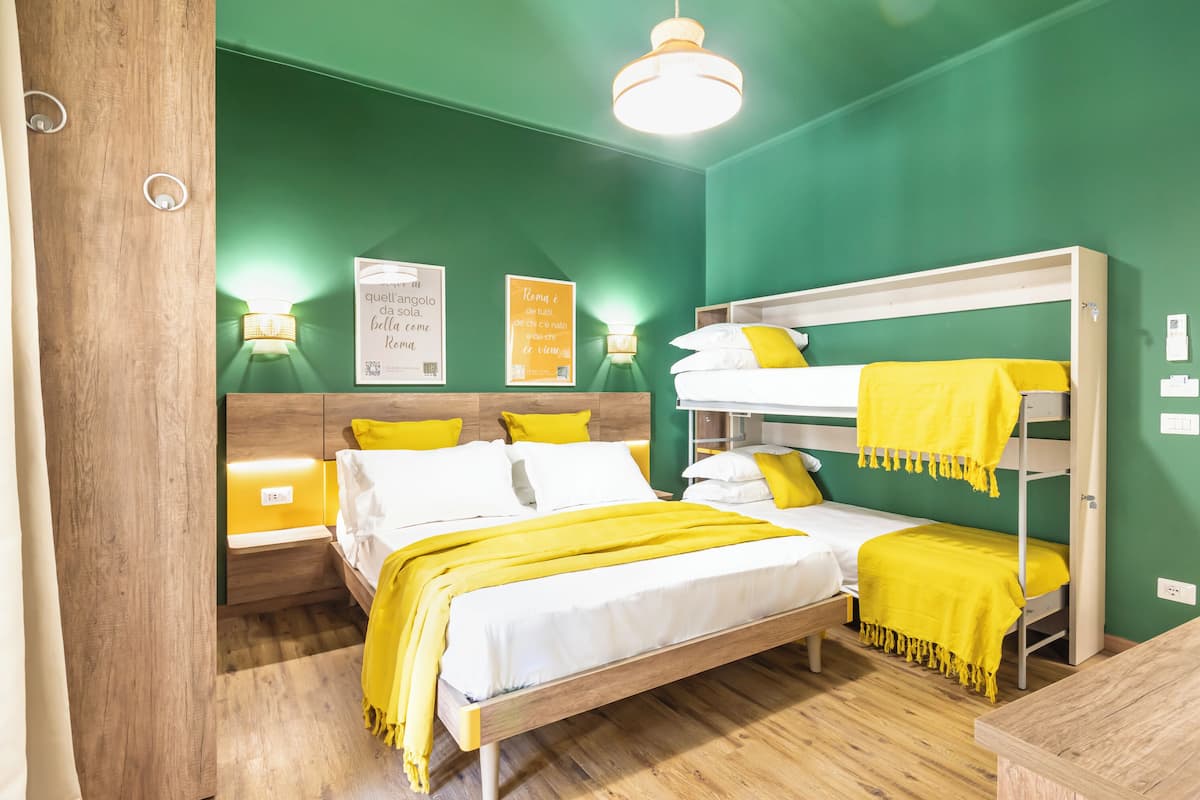 A room with a bunk bed, a single bed, and a wooden floor. There are two bedside tables with lamps, a green wall, and a ceiling light.