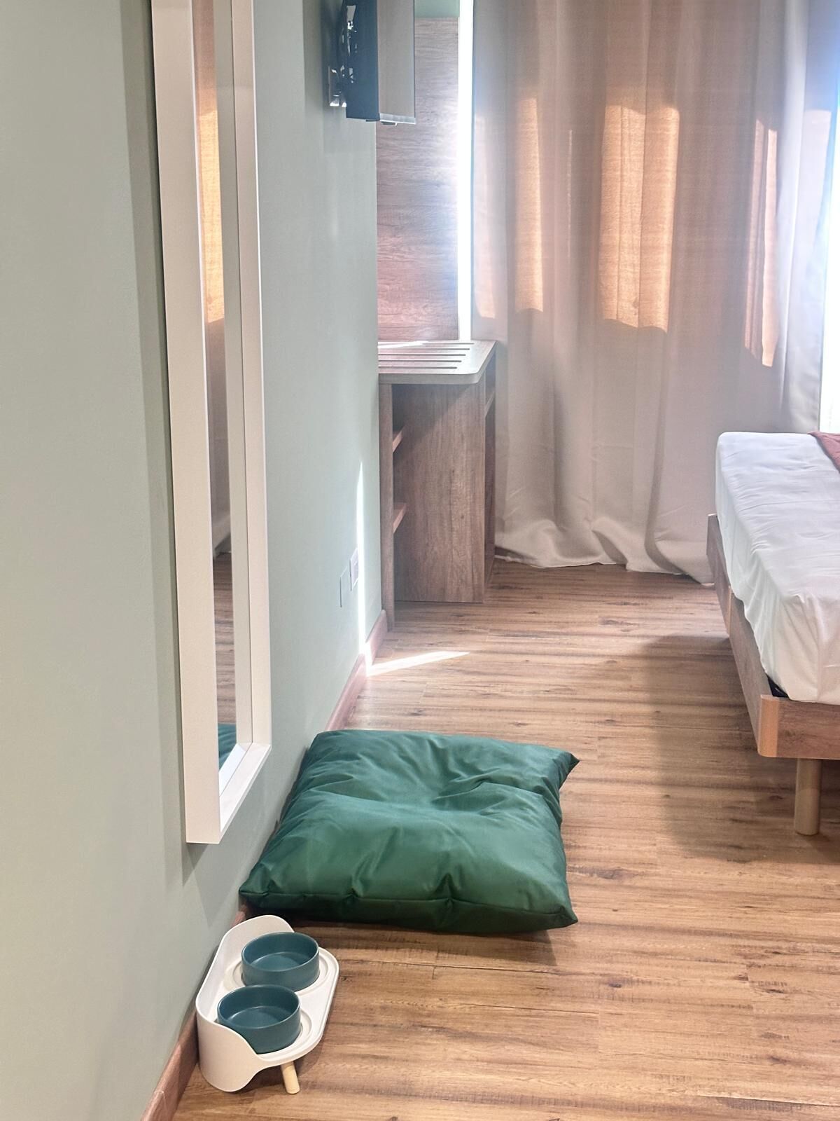 Una habitación estrecha con suelo de madera, una cama con ropa de cama blanca, un almoháriga verde y una cama para mascotas blanca con dos cuencos.